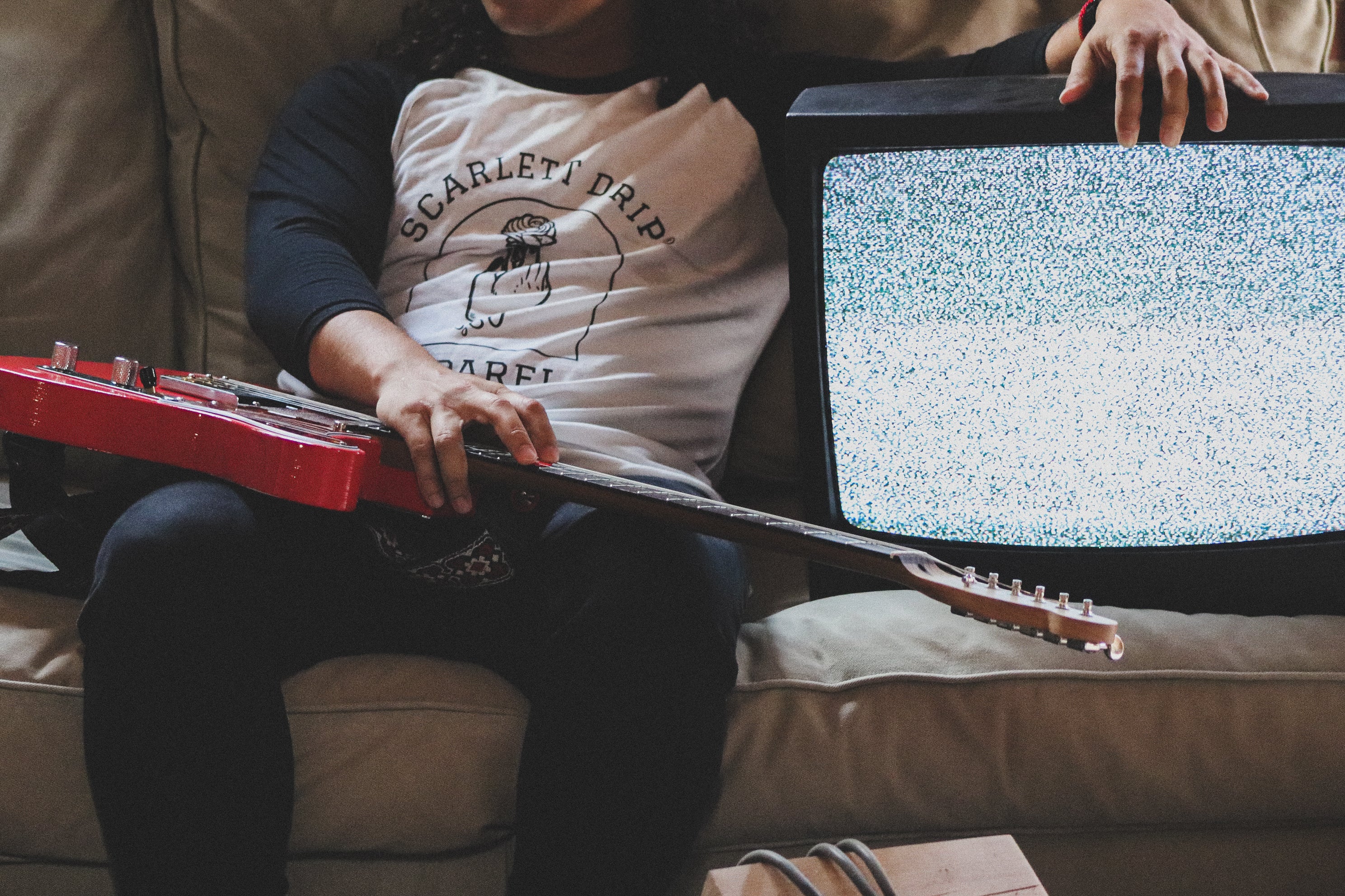 Person playing a red guitar in front of a vintage television set on a beige couch.